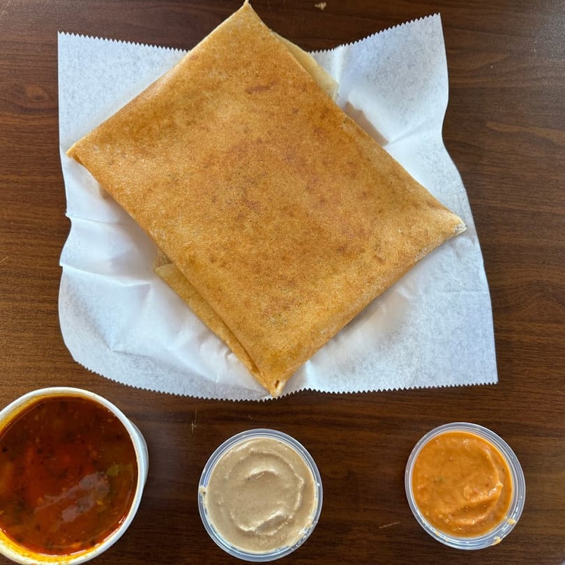 Avinya Indian Cuisine's dosa, folded on white paper, with three small sauce cups on a wooden table.